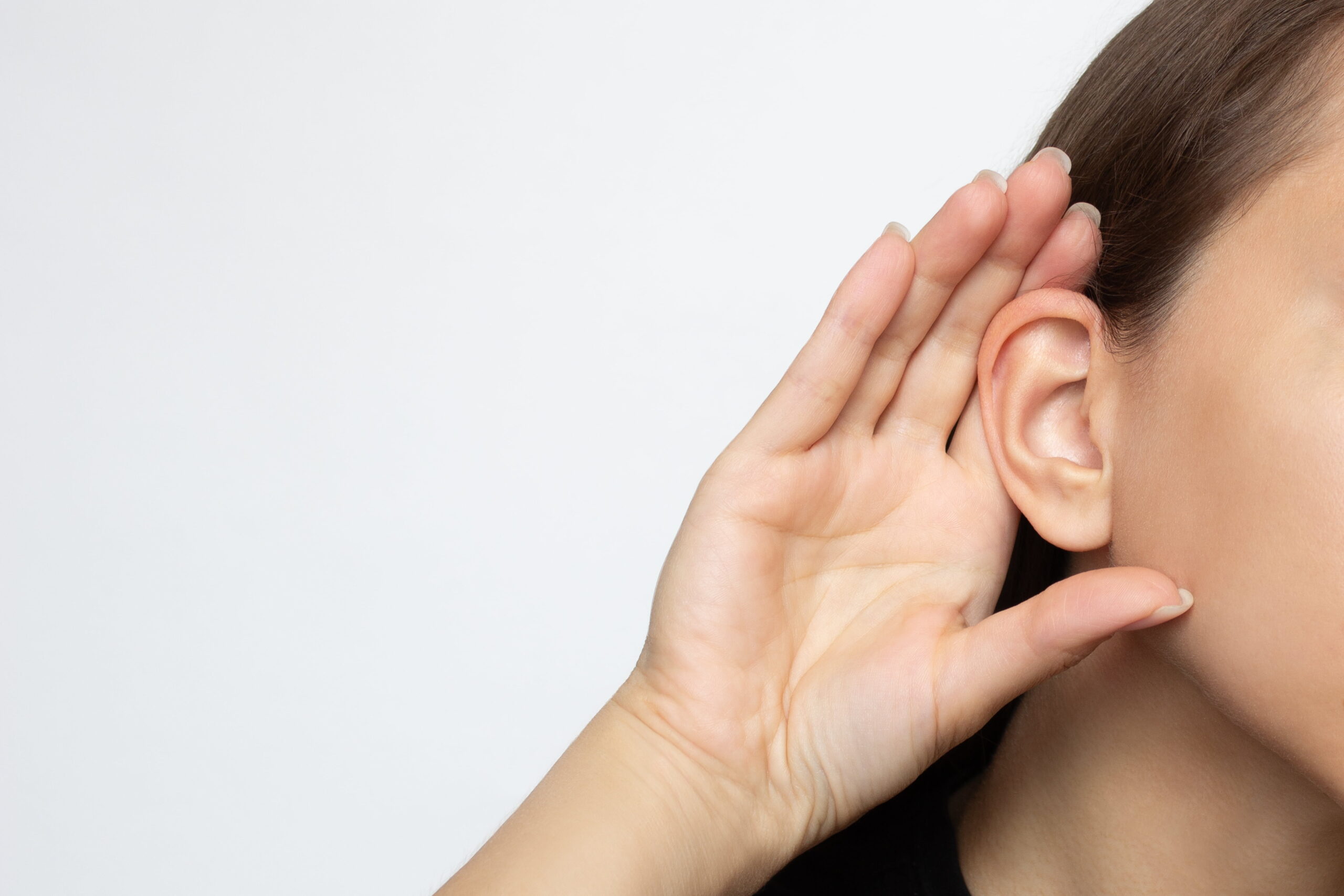 A person with long hair holds their hand to their ear, as if trying to hear better, against a plain light background. Only part of their face is visible.