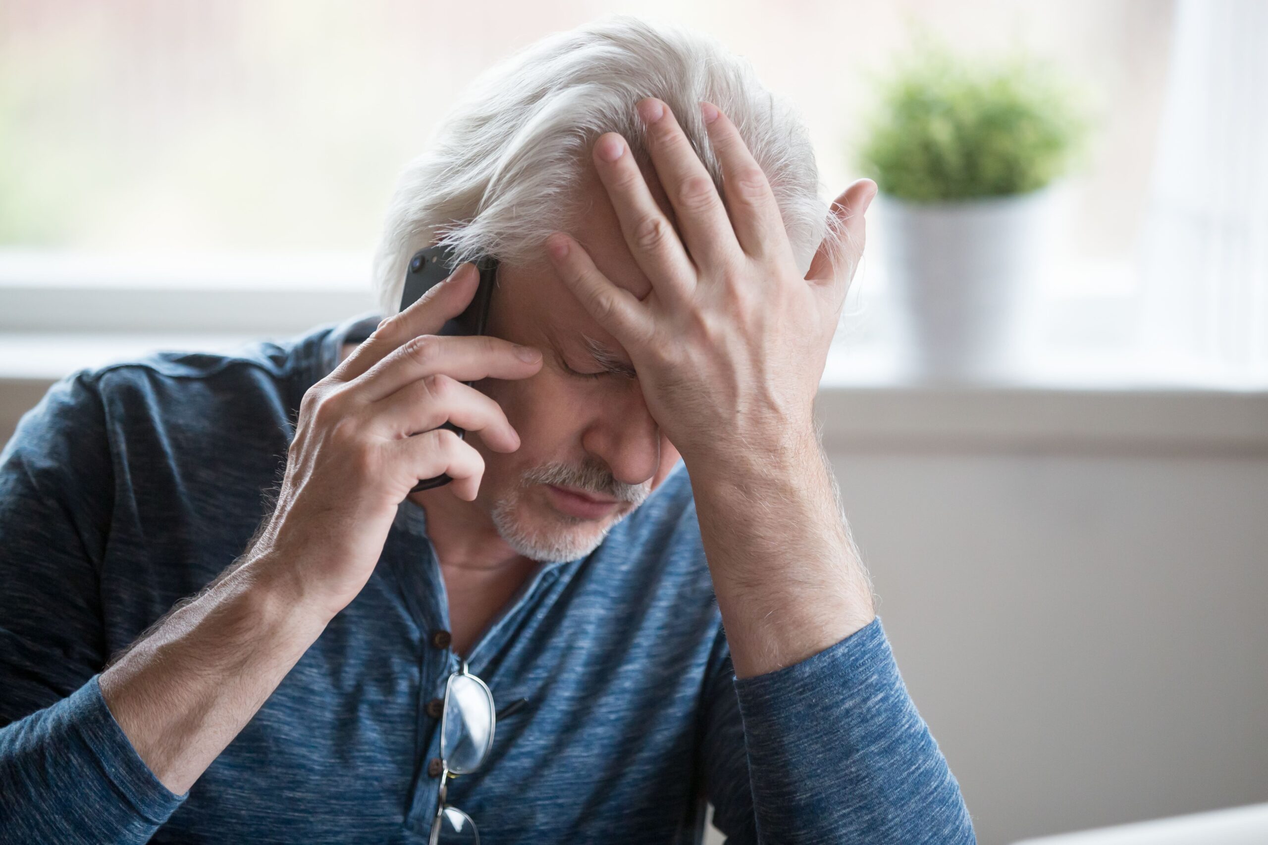 An older man with gray hair holds a cell phone to his ear and rests his other hand on his forehead, looking stressed or worried. He is wearing glasses and a blue shirt, sitting indoors with a plant in the background.
