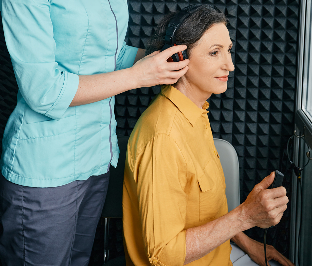 A woman in a yellow shirt sits in a soundproof room, wearing headphones, while another person in a teal uniform adjusts them. The woman holds a response button, likely for a hearing test.