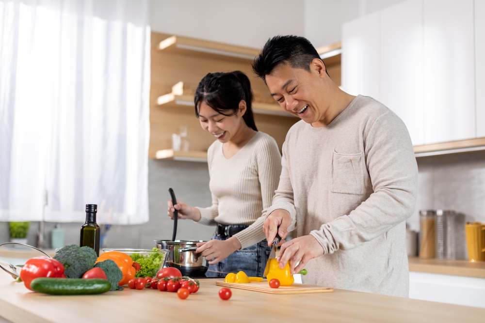 A smiling man slices a yellow bell pepper while a woman cooks at a stove in a bright kitchen. Fresh vegetables and cooking ingredients are spread out on the counter.