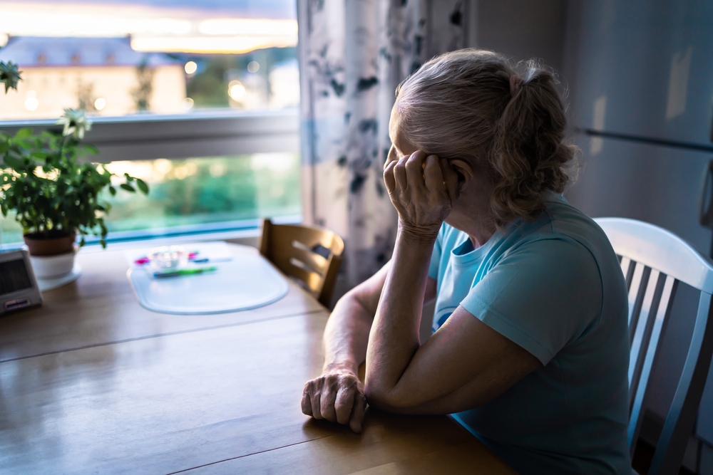 An elderly woman with gray hair sits at a kitchen table, resting her head on her hand and looking out the window, appearing thoughtful or lonely. Daylight filters through the window and a plant sits nearby.