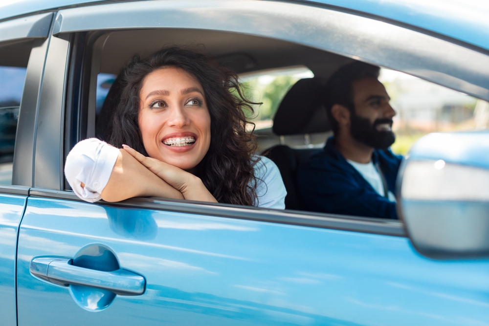 A smiling woman with braces leans her arms on the window of a blue car, looking outside. A bearded man is driving, both appear happy and relaxed.