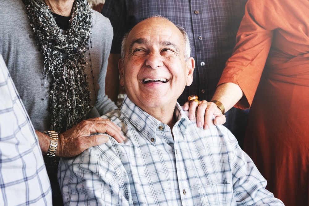 An older man wearing a plaid shirt smiles joyfully while seated, surrounded by friends who place their hands supportively on his shoulders. The group appears to be enjoying a warm, friendly moment together.