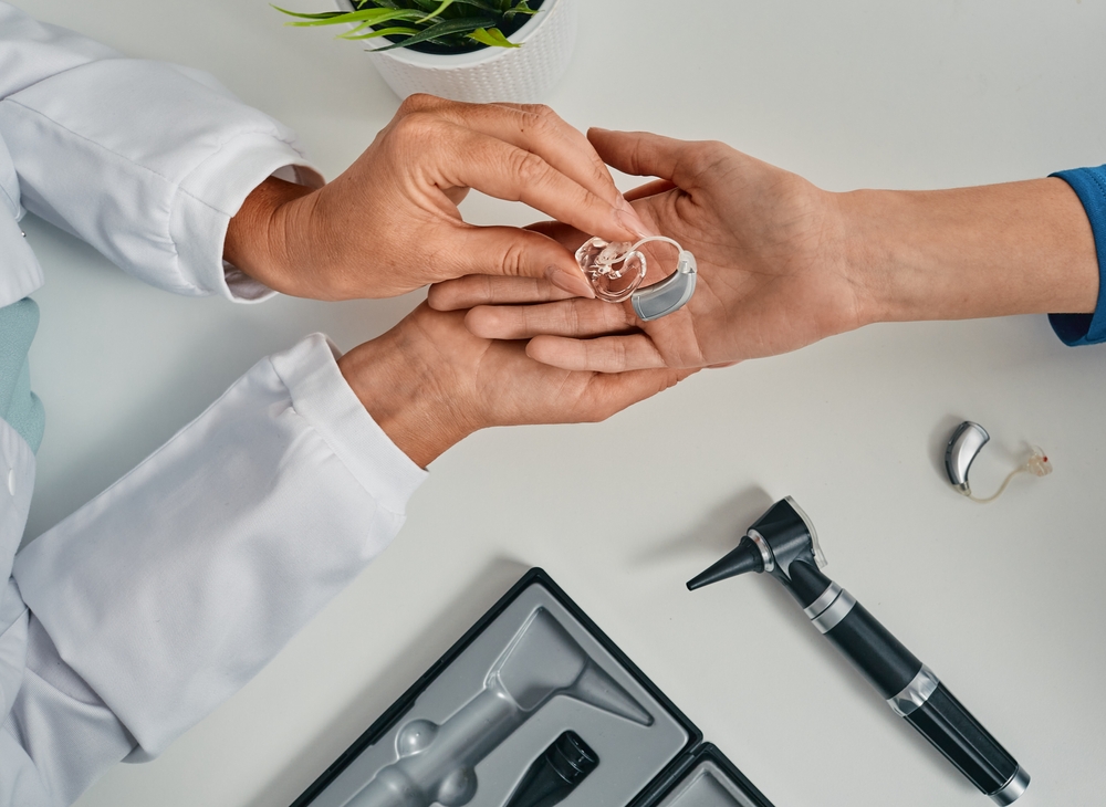 A doctor hands a hearing aid to a patient. Medical tools and a hearing aid case are on the table, along with a potted plant. Only their hands and arms are visible.