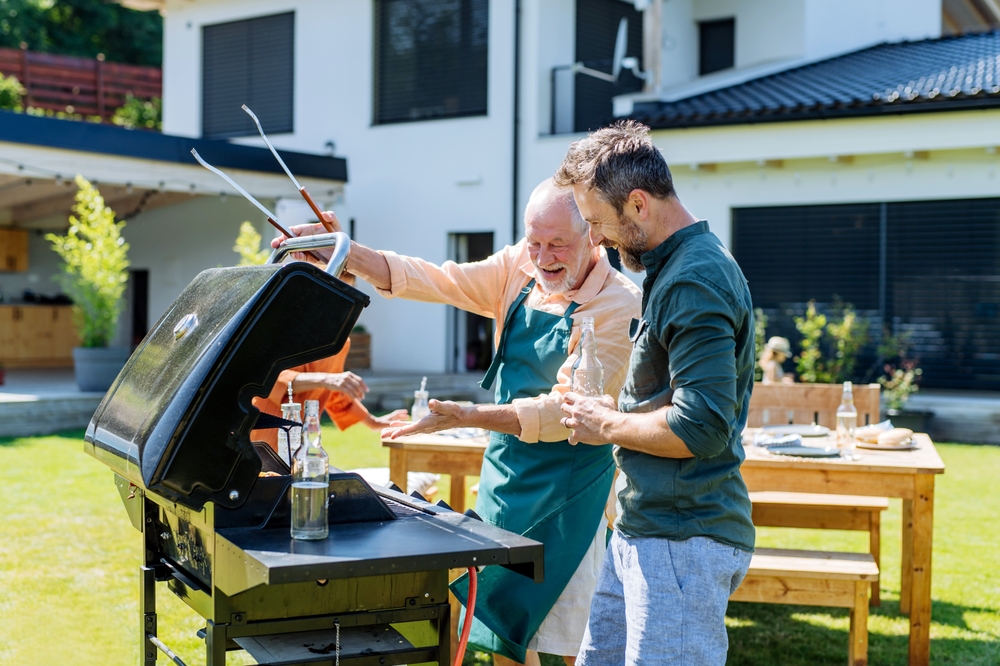 Two men stand by an outdoor grill in a backyard, smiling and grilling food together. One man wears an apron and gestures with tongs, while the other looks at the grill. A house and picnic table are in the background.