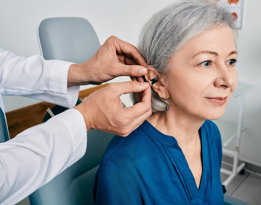 A doctor fits a hearing aid onto the ear of an older woman with gray hair, who is sitting in a medical office and wearing a blue shirt.