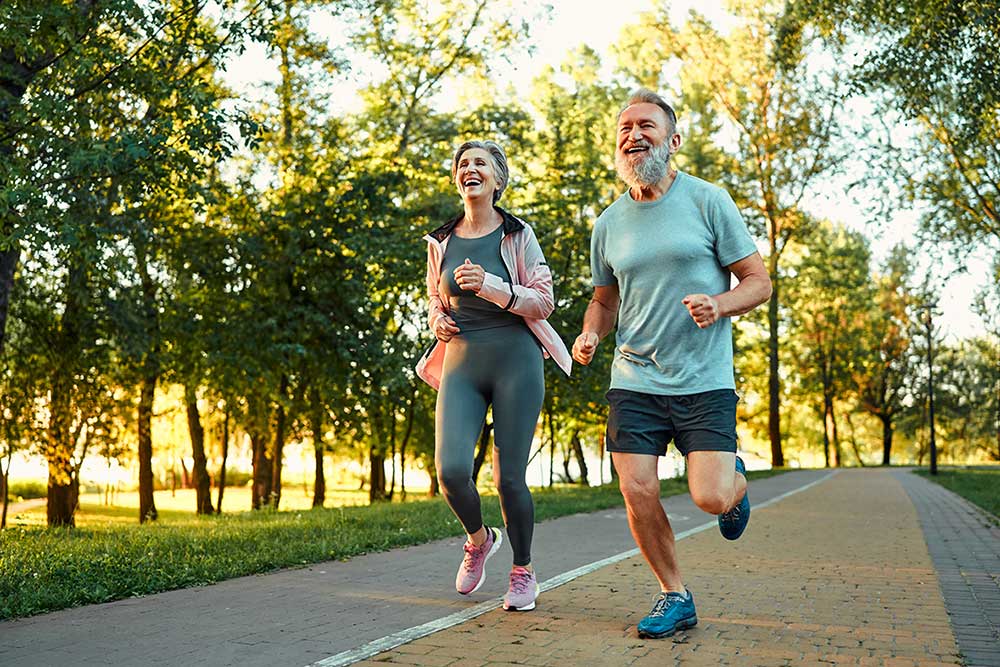 An older man and woman are jogging together on a paved park path, smiling and enjoying the outdoors. The sun is shining through trees, and both are dressed in athletic clothes.
