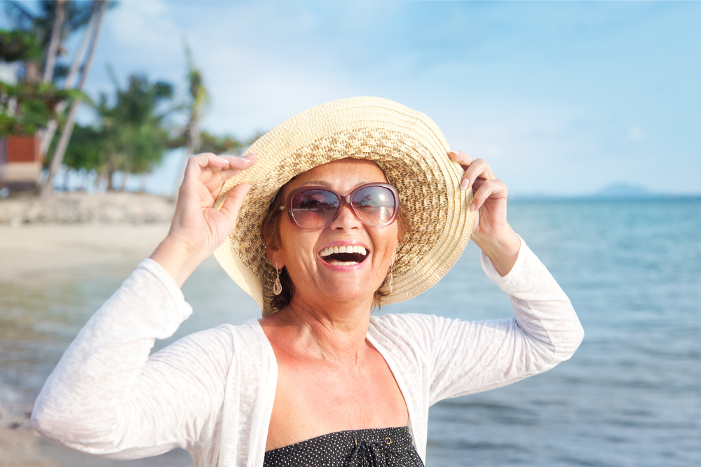 Smiling woman wearing sunglasses and a sunhat stands on a sunny beach near the ocean, with palm trees and blue sky in the background.