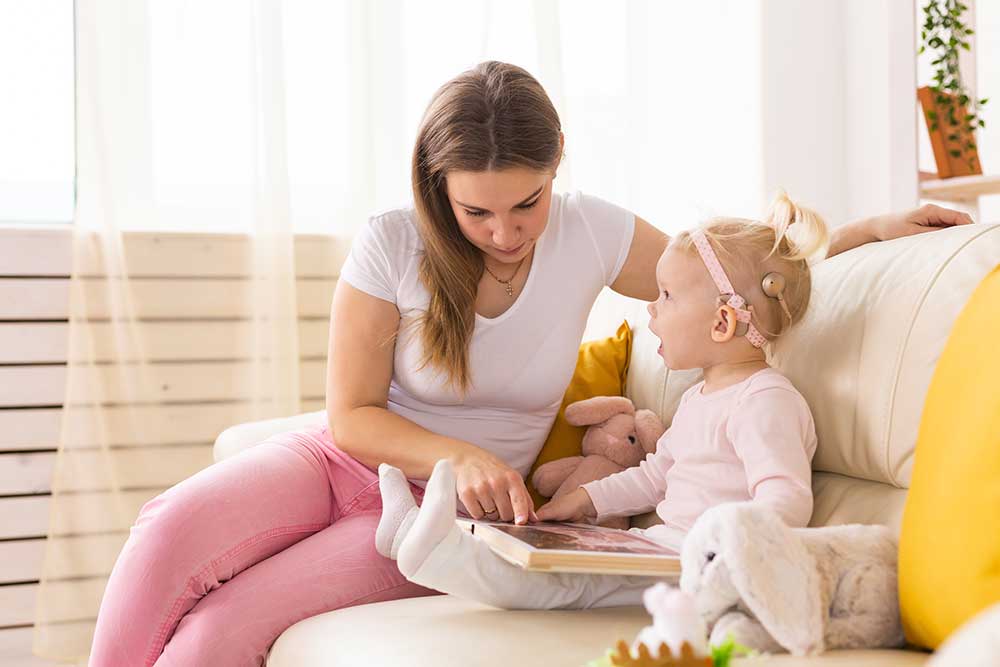 A woman sits on a couch with a young girl who has a cochlear implant. They are reading a book together, surrounded by soft toys, in a bright, cozy room.