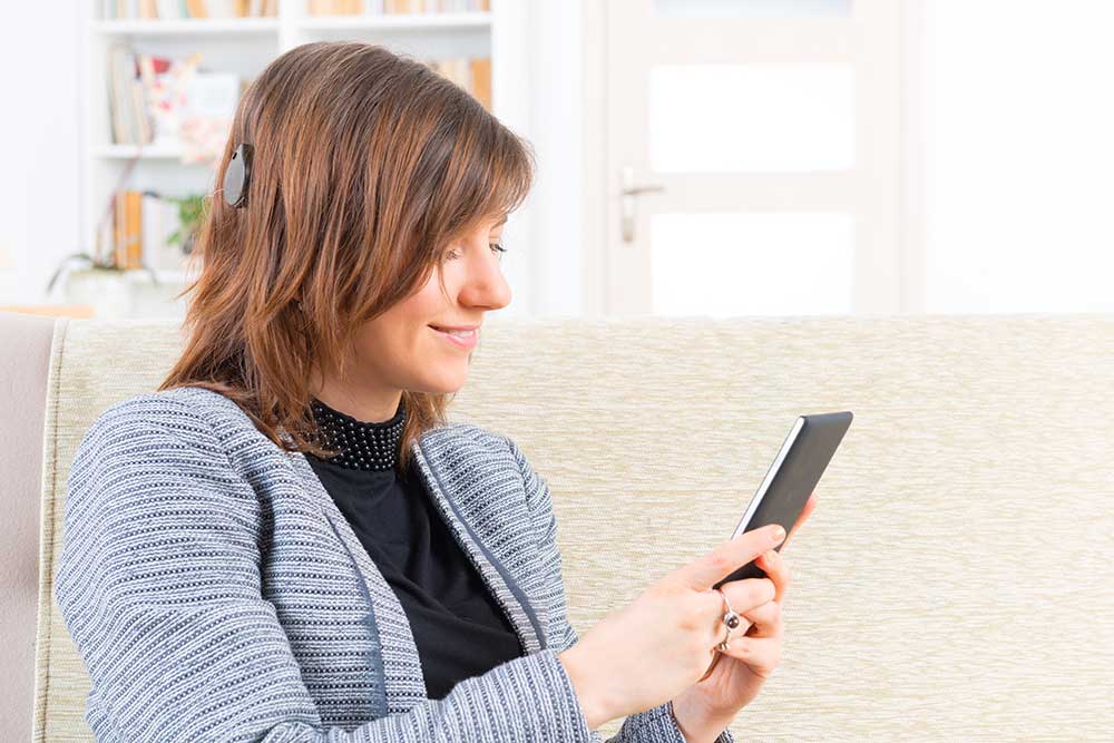 A woman with a cochlear implant sits on a sofa, smiling while using a smartphone. Shelves with books and a door are visible in the background.